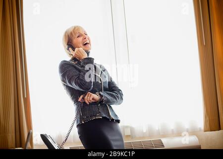 Die reife Geschäftsfrau lacht, als sie einen Anruf auf einem Festnetztelefon vor einem Fenster entgegennimmt. Stockfoto