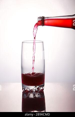Gießen Sie ein erfrischendes rotes, sprudelndes Sodagetränk aus der Flasche in Glas auf weißem Hintergrund mit Reflexionen Stockfoto