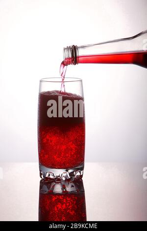 Gießen Sie ein erfrischendes rotes, sprudelndes Sodagetränk aus der Flasche in Glas auf weißem Hintergrund mit Reflexionen Stockfoto