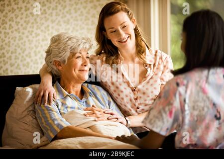 Junge Frau sitzt am Rand eines Bettes und legt einen beruhigenden Arm um eine Seniorin. Stockfoto