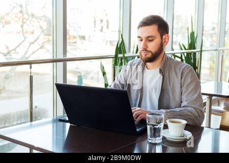 Remote-Arbeit. Geschäftsmann mit Laptop, kaukasischer Mann sitzt am Tisch im Café, arbeitet am Laptop, trinkt Kaffeewasser. Freiberufliche Arbeitsplätze, Mitarbeiter Stockfoto