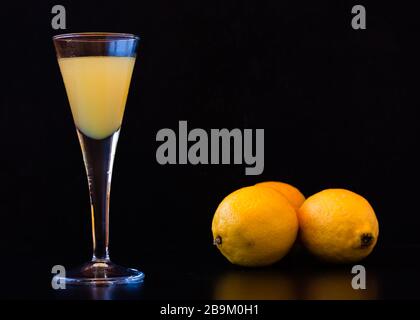 Limoncello in elegantem Schnapsglas mit drei Zitronen auf dunklem Hintergrund. Sizilianischer Zitronenlikör, aromatisch und erfrischend. Stockfoto