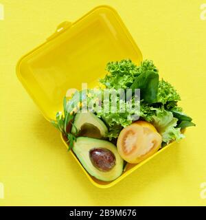 Lunchbox mit einem Snack aus Salat, Tomate, Avacado auf gelbem Hintergrund. Gesundes Esskonzept. Gehen Sie vegan. Leckere vegetarische Speisen in Kunststoffbox. Stockfoto