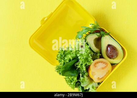 Lunchbox mit einem Snack aus Salat, Tomate, Avacado auf gelbem Hintergrund. Gesundes Esskonzept. Gehen Sie vegan. Leckere vegetarische Speisen in Kunststoffbox. Stockfoto