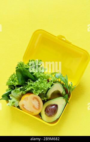 Lunchbox mit einem Snack aus Salat, Tomate, Avacado auf gelbem Hintergrund. Gesundes Esskonzept. Gehen Sie vegan. Leckere vegetarische Speisen in Kunststoffbox. Stockfoto