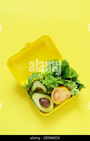 Lunchbox mit einem Snack aus Salat, Tomate, Avacado auf gelbem Hintergrund. Gesundes Esskonzept. Gehen Sie vegan. Leckere vegetarische Speisen in Kunststoffbox. Stockfoto