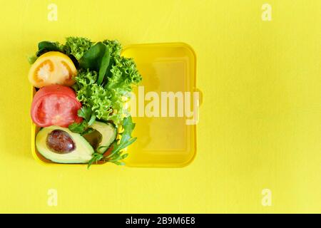 Lunchbox mit einem Snack aus Salat, Tomate, Avacado auf gelbem Hintergrund. Gesundes Esskonzept. Gehen Sie vegan. Leckere vegetarische Speisen in Kunststoffbox. Stockfoto