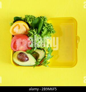 Lunchbox mit einem Snack aus Salat, Tomate, Avacado auf gelbem Hintergrund. Gesundes Esskonzept. Gehen Sie vegan. Leckere vegetarische Speisen in Kunststoffbox. Stockfoto