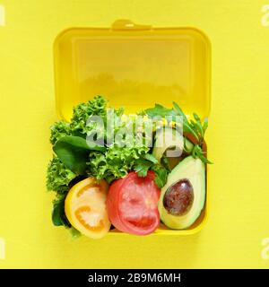 Lunchbox mit einem Snack aus Salat, Tomate, Avacado auf gelbem Hintergrund. Gesundes Esskonzept. Gehen Sie vegan. Leckere vegetarische Speisen in Kunststoffbox. Stockfoto