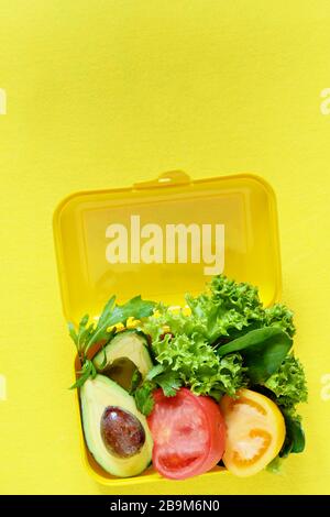 Lunchbox mit einem Snack aus Salat, Tomate, Avacado auf gelbem Hintergrund. Gesundes Esskonzept. Gehen Sie vegan. Leckere vegetarische Speisen in Kunststoffbox. Stockfoto