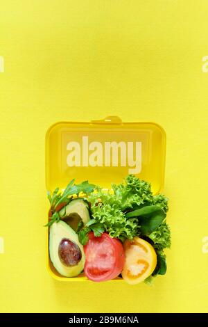 Lunchbox mit einem Snack aus Salat, Tomate, Avacado auf gelbem Hintergrund. Gesundes Esskonzept. Gehen Sie vegan. Leckere vegetarische Speisen in Kunststoffbox. Stockfoto