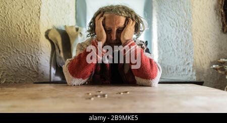 Verzweifelter älterer Mann mit Tränen in den Augen, der mit ein paar Münzen auf seinem bescheidenen Schreibtisch sitzt. Konzeptionelles Bild der Armut. Stockfoto