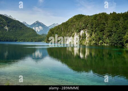 Alpsee bei Hoehenschwangau und Schloss Neuschwanstein in Bayern, Deutschland. Klarer Sommerhimmel und Alpen im Hintergrund. Stockfoto