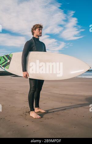 Junger Surfer, der mit seinem Surfbrett in einem schwarzen Surfanzug im Ozean steht. Sport- und Wassersportkonzept. Stockfoto