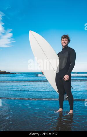 Junger Surfer, der mit seinem Surfbrett in einem schwarzen Surfanzug im Ozean steht. Sport- und Wassersportkonzept. Stockfoto