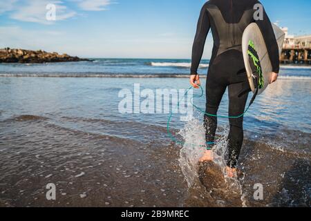 Junger Surfer, der mit seinem Surfbrett in einem schwarzen Surfanzug ins Wasser tritt. Sport- und Wassersportkonzept. Stockfoto