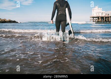 Junger Surfer, der mit seinem Surfbrett in einem schwarzen Surfanzug ins Wasser tritt. Sport- und Wassersportkonzept. Stockfoto