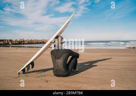 Junger Surfer sitzt am Sandstrand und neben seinem Surfbrett. Sport- und Wassersportkonzept. Stockfoto