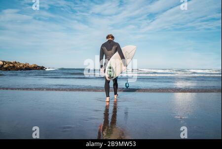 Junger Surfer, der mit seinem Surfbrett in einem schwarzen Surfanzug ins Wasser tritt. Sport- und Wassersportkonzept. Stockfoto