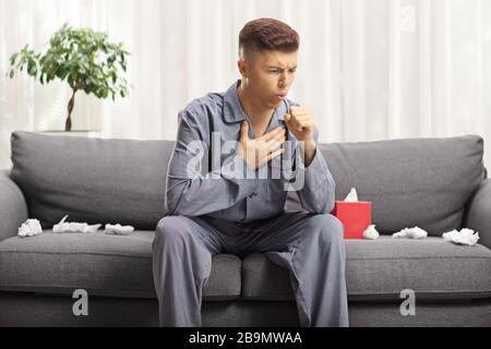 Junger Mann hustet, fühlt sich krank und sitzt auf einem Sofa im Schlafanzug mit Gewebepapieren zu Hause Stockfoto