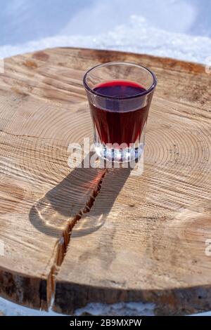 Alkoholhaltige rote Tinktur in einem kleinen Glas steht auf einem geschnittenen Baumstumpf. Schnaps in einem Glas. Konzentrieren Sie sich auf das Glas. Vertikal. Stockfoto