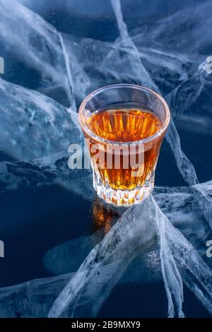 Whisky in einem kleinen Glas steht auf dem transparenten Eis des Baikalsee. Tiefe schöne Risse im Eis. Konzentrieren Sie sich auf ein Glas Alkohol. Vertikal. Stockfoto