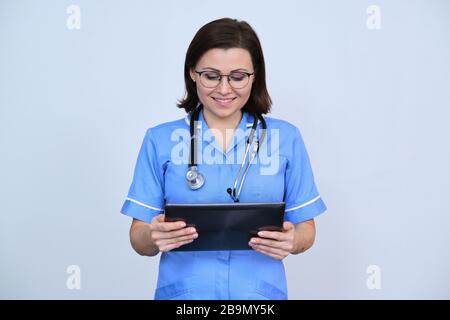Mittelalter-Ärztin mit digitaler Tablette, lächelnde Krankenschwester, die auf Tablette blickt, liest, auf grauem Hintergrund Stockfoto