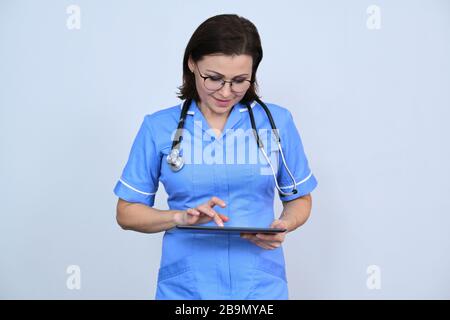 Mittelalter-Ärztin mit digitaler Tablette, lächelnde Krankenschwester, die auf Tablette blickt, liest, auf grauem Hintergrund Stockfoto