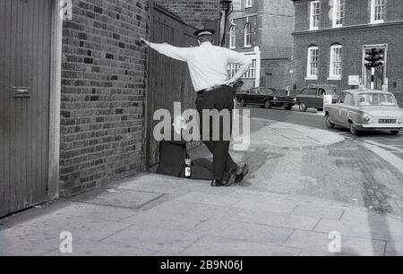 1973, historisch, ein britischer Polizist oder "bobbie" stehend, der mit einem Mann spricht, der auf dem Straßenbelag sitzt, der mit einer Flasche Getränk in der Hand schlummert, London, England, Großbritannien. In dieser Epoche und in den letzten Jahrzehnten würde der lokale "Bobbie" sein Territorium betreten oder patrouillieren oder die Umgebung und die Einheimischen schlagen und kennen. Diese traditionelle Polizeiform, die auf einer engen Beziehung zur Gemeinde basierte, veränderte sich in späteren Jahrzehnten, da die Polizei zu einer Truppe wurde, die aus Autos und nicht zu Fuß operierte. Stockfoto