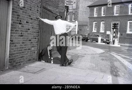 1973, historisch, ein britischer Polizist oder "bobbie" stehend, der mit einem Mann spricht, der auf dem Straßenbelag sitzt, der mit einer Flasche Getränk in der Hand schlummert, London, England, Großbritannien. In dieser Epoche und in den letzten Jahrzehnten würde der lokale "Bobbie" sein Territorium betreten oder patrouillieren oder die Umgebung und die Einheimischen schlagen und kennen. Diese traditionelle Polizeiform, die auf einer engen Beziehung zur Gemeinde basierte, veränderte sich in späteren Jahrzehnten, da die Polizei zu einer Truppe wurde, die aus Autos und nicht zu Fuß operierte. Stockfoto