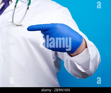 medic man in weißem Mantel mit Knöpfen, an Händen mit blauen sterilen Handschuhen, zeigt Handgeste, die auf das Motiv hinweist, blauer Hintergrund Stockfoto