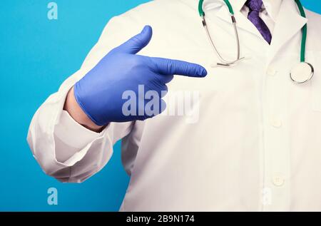 medic man in weißem Mantel mit Knöpfen, an Händen mit blauen sterilen Handschuhen, zeigt Handgeste, die auf das Motiv hinweist, blauer Hintergrund Stockfoto
