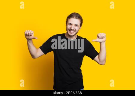 Selbstbewusster kaukasischer Mann mit Bart und langem Haar, der auf ihn zeigt, während er auf einem gelben Hintergrund posiert Stockfoto