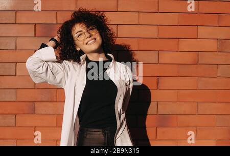 Geschweifte kaukasische Frau, die draußen auf einer Steinwand posiert, während sie Brille und ein schönes weißes Hemd trägt Stockfoto