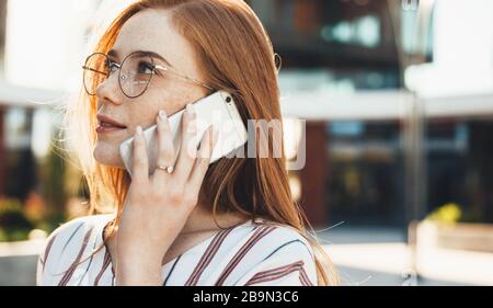 Frecklige kaukasische Unternehmerin mit roten Haaren und Brillen hat draußen eine Telefondiskussion Stockfoto