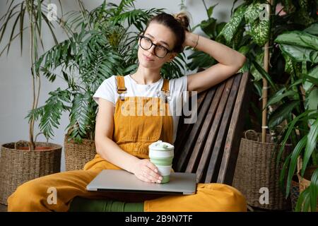 Weibliche europäische Gärtnerin, die Overalls trägt, nach der Arbeit ruht, auf Holzstuhl im heimischen Gewächshaus sitzt, wiederverwendbaren Kaffee-/Teebecher beiseite hält Stockfoto