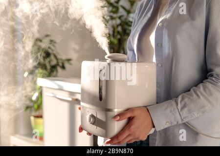 Frau, die in ihren Händen arbeitet, arbeitet mit Aromaölbefeuchter im sonnigen Raum. Erhöhung der Luftfeuchtigkeit im Innenbereich, komfortable Lebensbedingungen in der Heizung Stockfoto