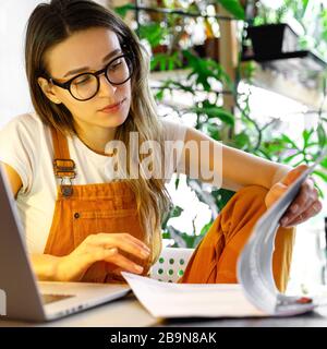 Junge Gärtnerin in Brille mit Laptop, erstellt einen Bericht über Bürodokumente, arbeitet von zu Hause aus während der Quarantäne wegen Coronavirus. Gemütliches Büro mit Stockfoto