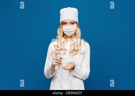Weibliche Ärztin, die eine chirurgische Gesichtsmaske trägt, die Desinfektionsmittel mit Alkoholgel hält, isoliert auf blauem Grund. Junge Frau in medizinischer Uniform und Schutzkleidung Stockfoto