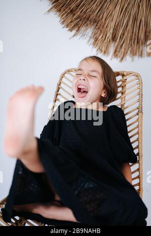 Wildes, frech kleines Mädchen in einem schwarzen Kleid, das auf einem Stuhl sitzt und schreit. Stockfoto