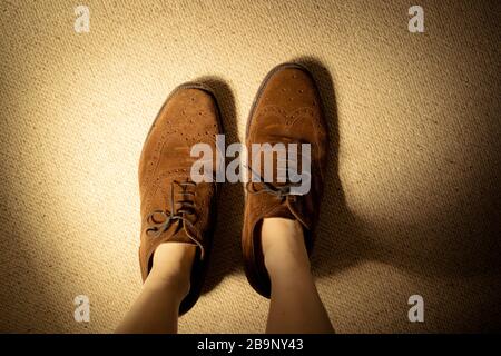 Blick hinunter auf die Füße einer Frau oder eines Kindes, das braune Wildleder-Brogue-Schuhe eines großen Mannes trägt, die viel zu groß sind. Stockfoto