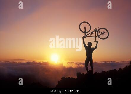 Mann hält Fahrrad bei Sonnenaufgang am Haleakala Krater, Maui, Hawaii über Kopf. Stockfoto