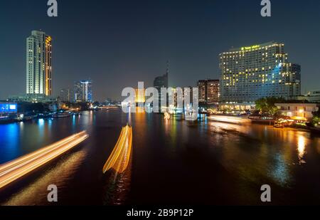 Langzeitfotografie des Chao-Phraya-Flusses mit verschwommenen Bewegungen beleuchteter Boote und der städtischen Skyline von Bangkok in der Nacht, Thailand. Stockfoto