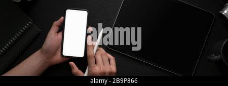 Overhead-Aufnahme eines Mannes, der ein leeres Bildschirm-Smartphone in einem dunklen, modernen Arbeitsbereich mit anderen Büromaterialien hält Stockfoto