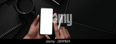 Overhead-Aufnahme eines Mannes, der ein leeres Bildschirm-Smartphone in einem dunklen, modernen Arbeitsbereich mit anderen Büromaterialien hält Stockfoto