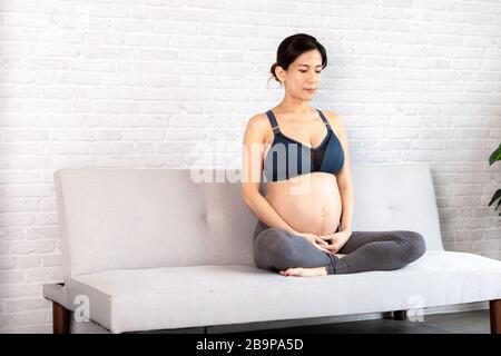 Ganzkörperschwangerin Chinesin Asian in Sportswear sitzend gekreuzt auf der Couch und meditierend während des Yoga-Trainings im Wohnzimmer Stockfoto