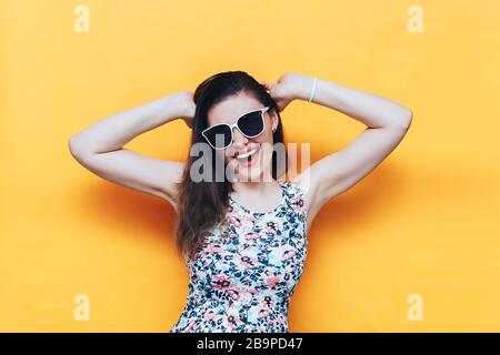 Fröhliche schöne lachende junge Frau im Kleid und weiße Sonnenbrille auf gelbem Hintergrund Stockfoto