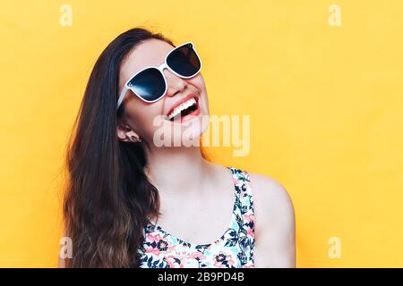 Fröhliche schöne lachende junge Frau im Kleid und weiße Sonnenbrille auf gelbem Hintergrund Stockfoto