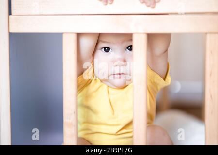 Süßes Baby, das im Kinderbett sitzt und durch den Holzrahmen über das Zimmer blickt. Sie hat Angst vor etwas. Baby versteckt sich in der Krippe Stockfoto