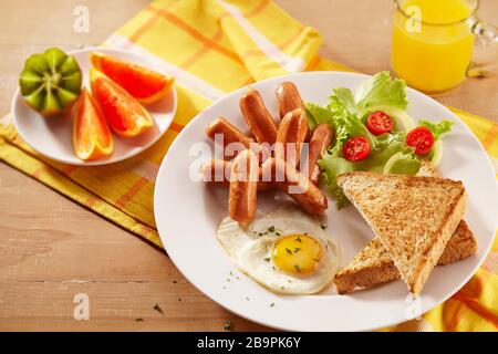 Ein Frühstücksplatte mit Cocktailwürsten, geröstetem Brot, Spiegeleiern, Salaten, Obst und einem Glas Orangensaft Stockfoto
